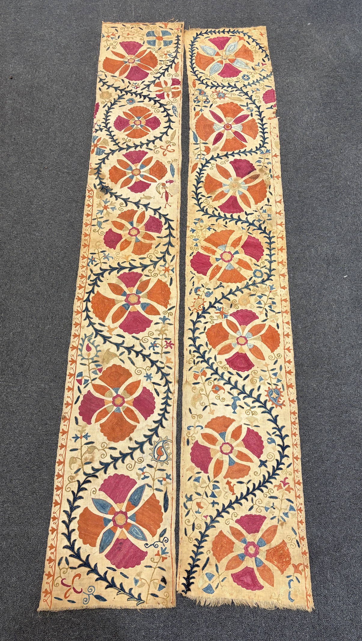A pair of Uzbekistan long wide panels, both embroidered in mostly red, coral and blue with large symbolic flowers and trailing vines on woven cotton using mostly Burkara couching, longest 250cm long x 38cm wide